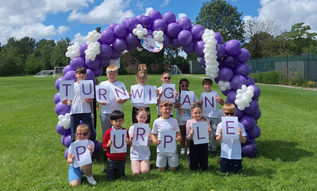 A group of smiling children under a purple and white balloon arch holding letters that read: "Turn Wigan Purple"